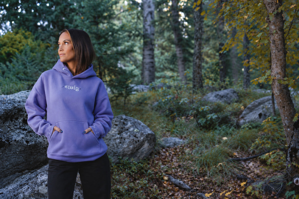 Woman wearing the purple hoodie in a woodland setting, with her hands tucked into the front pocket.