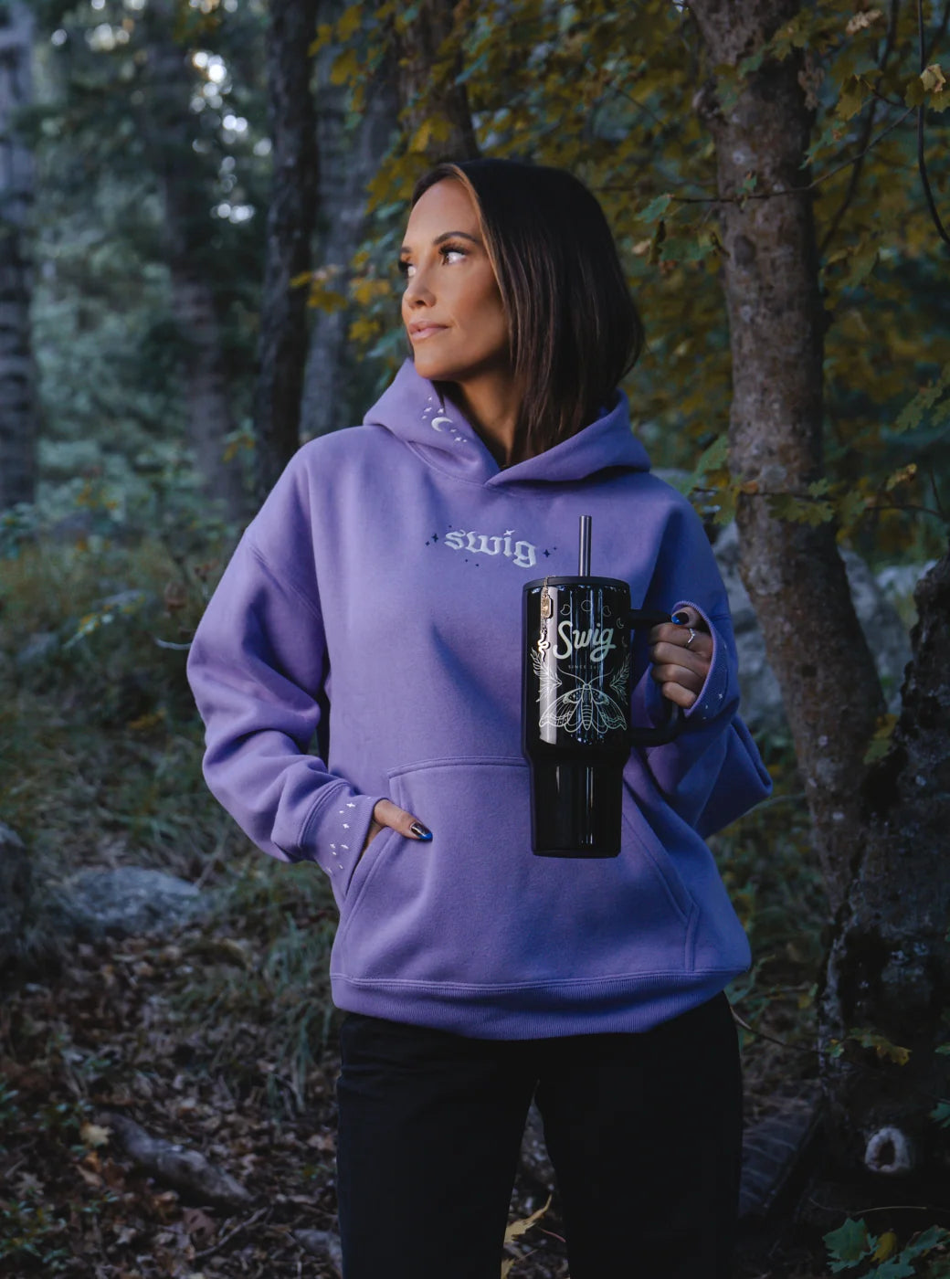 Portrait of a model in a lavender Swig hoodie holding a black moth tumbler in the woods.