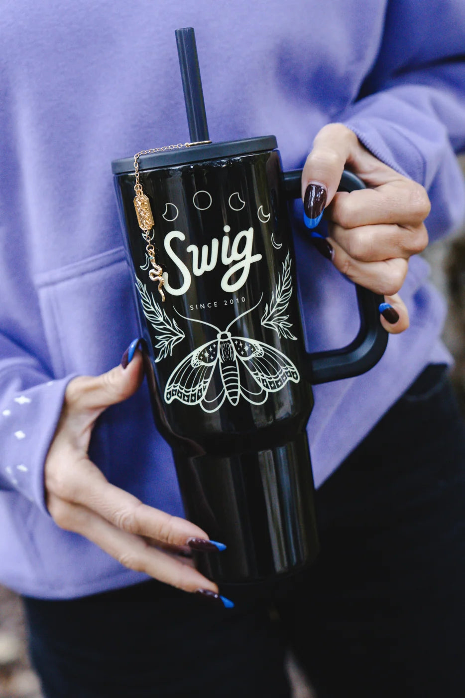 Close-up of a black moth tumbler held in two hands against a lavender Swig hoodie.
