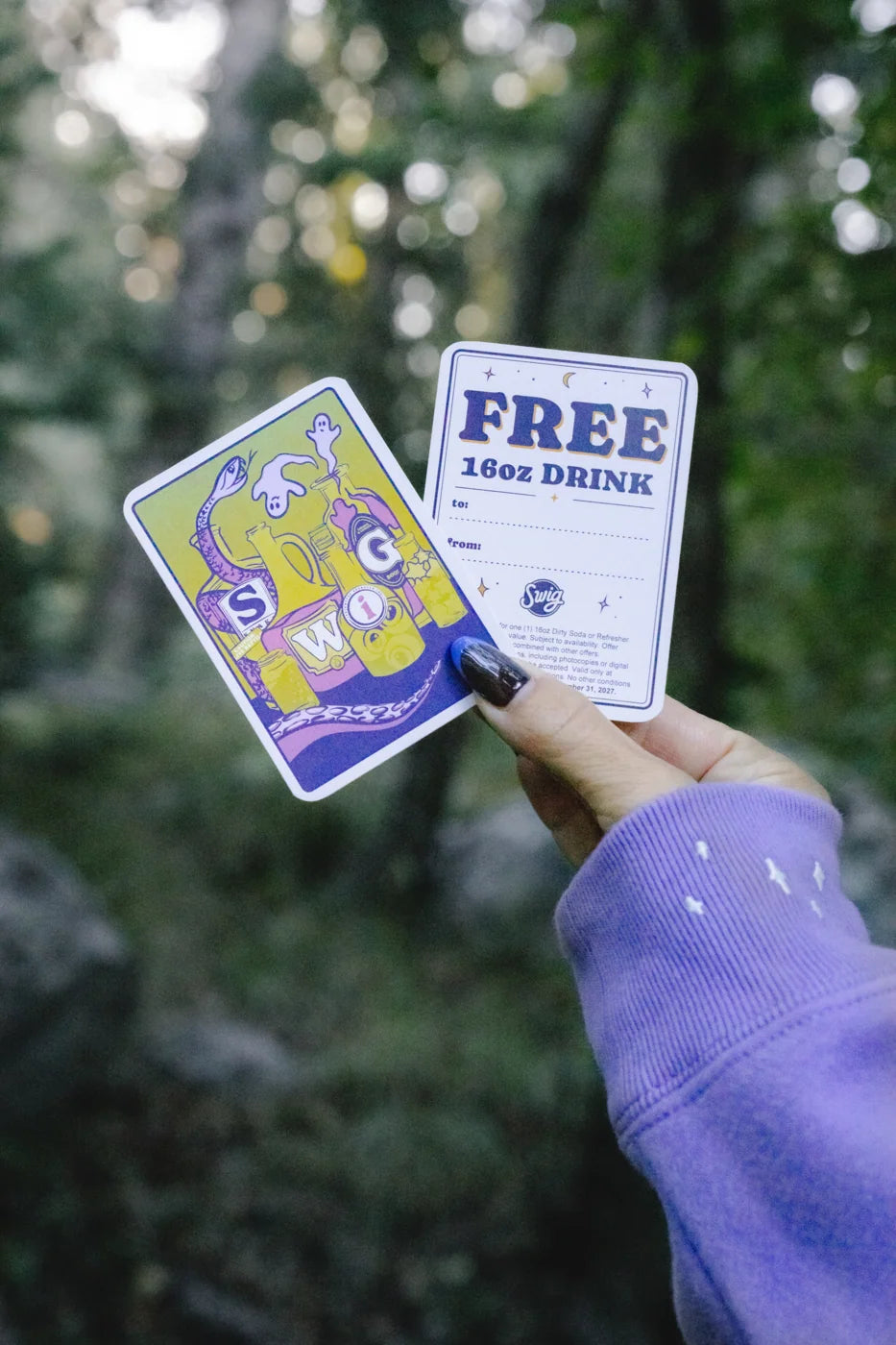 A hand in a lavender sleeve holds two sides of a Swig illustrated card, including a free 16 oz drink message.