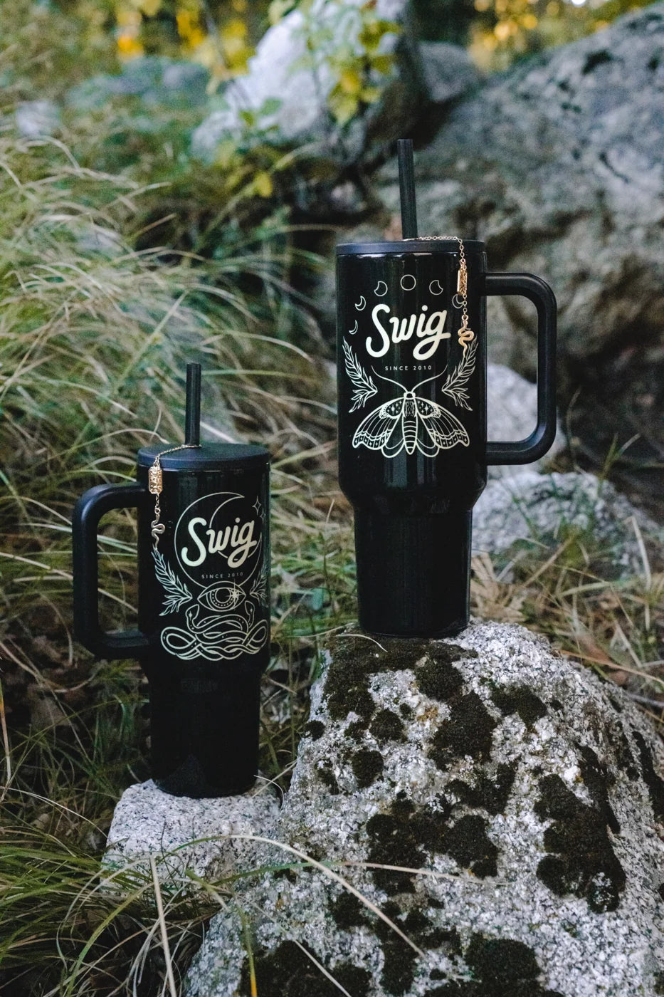 Two black illustrated Swig tumblers set at different heights on rocks beside woodland plants.